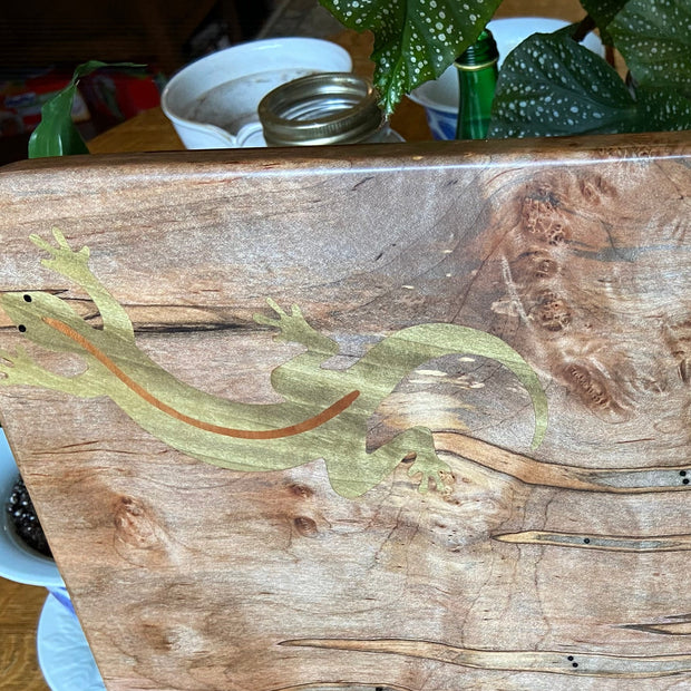 Wooden cutting board with a stand on a wooden surface, surrounded by plants.